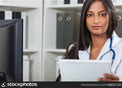 An Indian Asian female medical doctor in a hospital office with stethoscope using tablet computer&#xD;