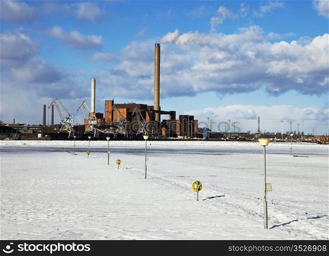 An industrial facility in Helsinki is emitting steam from a smokestack. In winter, the harbor is filled with ice and the marina in the foreground is empty.