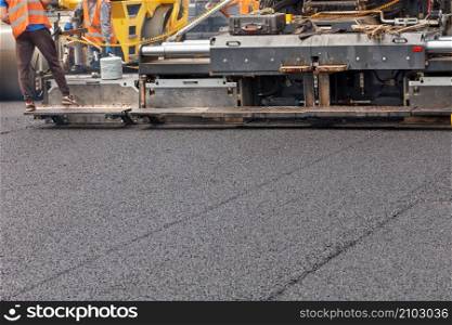 An industrial paver is laying a layer of new hot asphalt on the roadway. Workers control the evenness and height of the paving. Copy space.. Construction of a new road, laying of fresh asphalt with industrial road equipment.