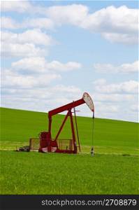 An oil well with the pump jack in action, against a grassy, green hill & cloudy blue sky. Located in the province of Alberta, Canada.