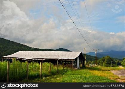 An old barn shed at the side of a road in a rural area with mountains and sky in the distance.
