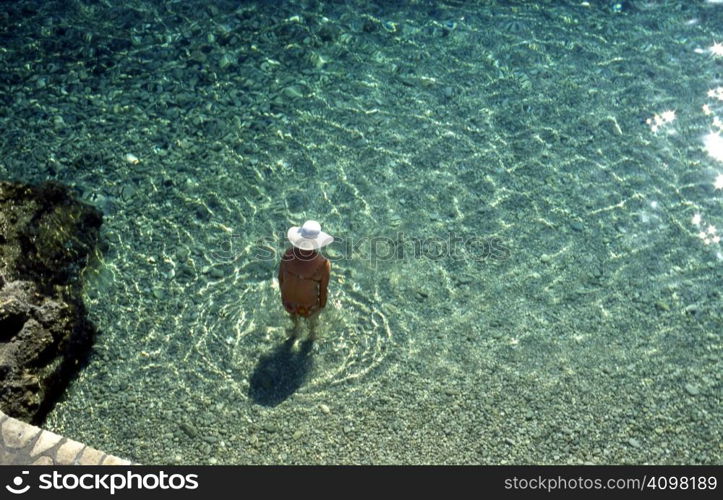 An old lady in a wide brimmed hat, paddling in shallow water in a cove on the island of Ithaca, Greece