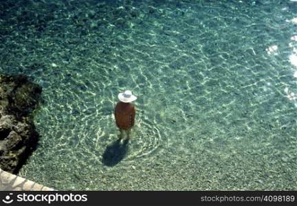 An old lady in a wide brimmed hat, paddling in shallow water in a cove on the island of Ithaca, Greece