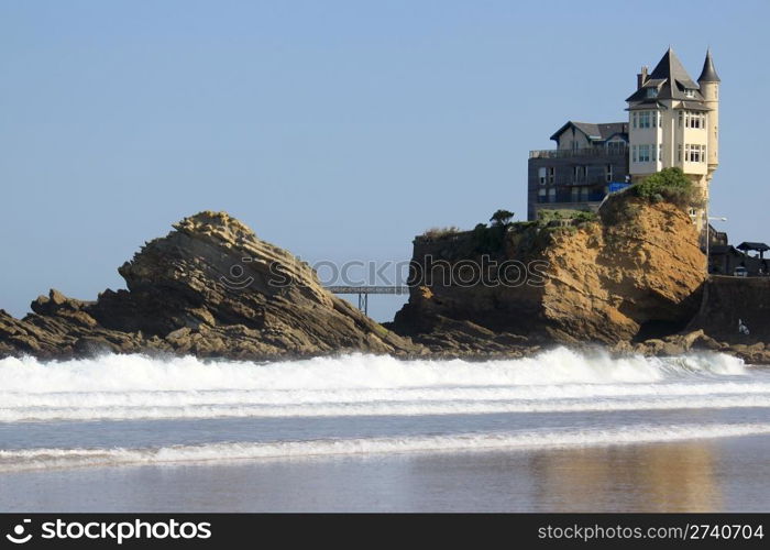 An old mansion perched on a rock in the ocean near a beach on a blue sky background