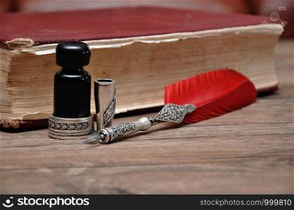 An old pen with a bottle of ink on a wooden table with a red book