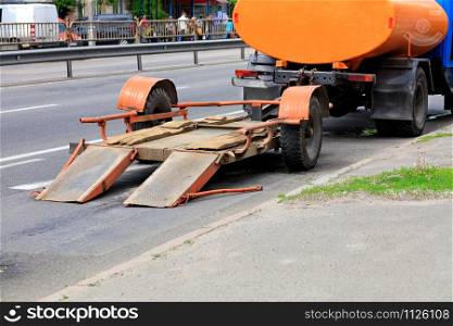 An old trailer with a low platform for transporting road equipment is attached to the rear bumper of an old water carrier truck, image with copy space.. An old low-platform trailer hitched to an old truck that stands on a roadway.
