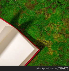 An open book with a red cover lies on the green grass or moss. View from above. book in grass