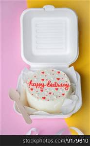 An open box with a birthday bento cake stands on a yellow background. A small bento birthday cake in a box with a wooden spoon. Yellow and pink background
