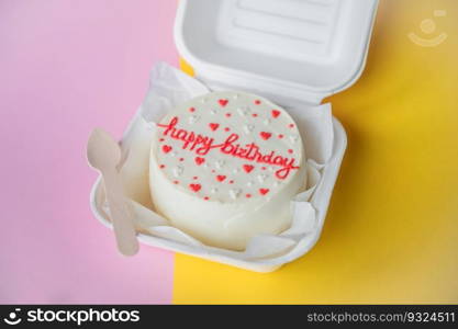 An open box with a birthday bento cake stands on a yellow background. A small bento birthday cake in a box. Wooden boat with white ribbon