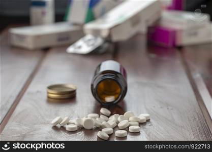 an open glass pill bottle with round white pills. On the background of different boxes with pills and blisters. Front view, wooden background. an open glass pill bottle with round white pills. On the back of different boxes with pills and blisters.