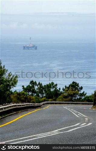 An open road at a mountain pass lies in front of the ocean with an oil drill platform in the far distance.
