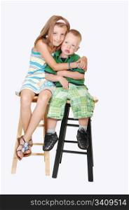 An pretty girl sitting with her brother on chairs and giving him a big hugon light blue background.