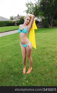 An slim young girl in a turquoise bikini and an yellow scarf in her hand dancing on the grass in the backyard by the beach.