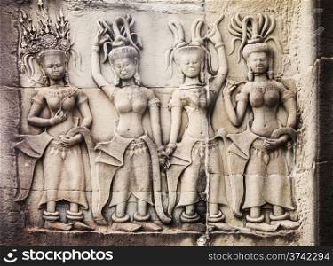 Ancient stone carvings on a wall at Angkor Wat in Cambodia depict four women standing together. They are attired similarly but are wearing fancy hats.