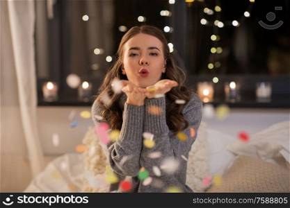 and people concept - young woman blowing colorful confetti from her hands at home. woman blowing confetti from her hands at home