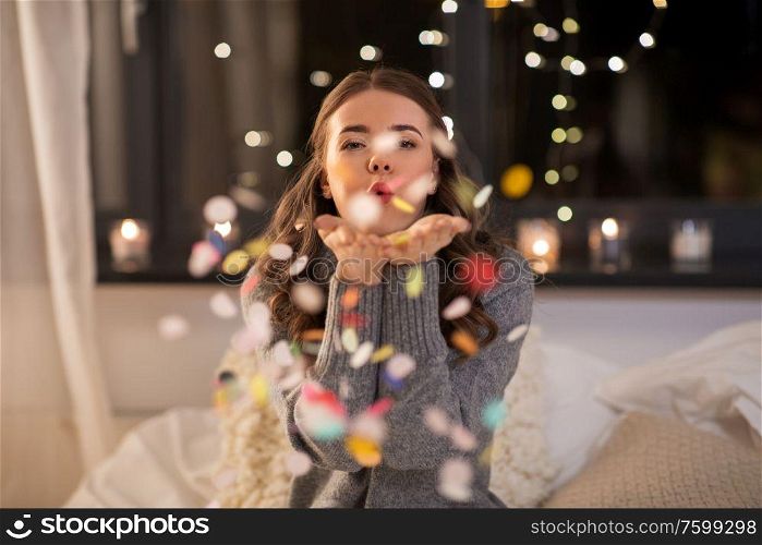 and people concept - young woman blowing colorful confetti from her hands at home. woman blowing confetti from her hands at home