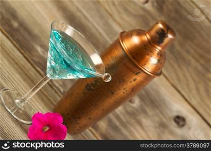 Angled horizontal view of a mixed drink, bright pink flower, stir stick and a metal mixer resting on rustic wood