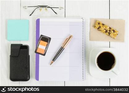 Angled top view image of a business desktop consisting of the following items: calendar, pen, coffee, cell phone and case, reading glasses, snack, napkin, and paper notes on white wood.