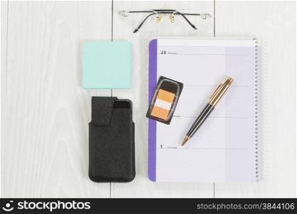 Angled top view image of a business desktop consisting of the following items: calendar, pen, reading glasses, paper stickers and cell phone inside of case on white wood.