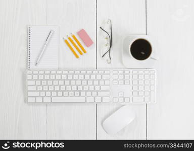 Angled top view image of a business desktop consisting of the following items: keyboard, mouse, pen, pencils, coffee, reading glasses, and note pad on white wood.