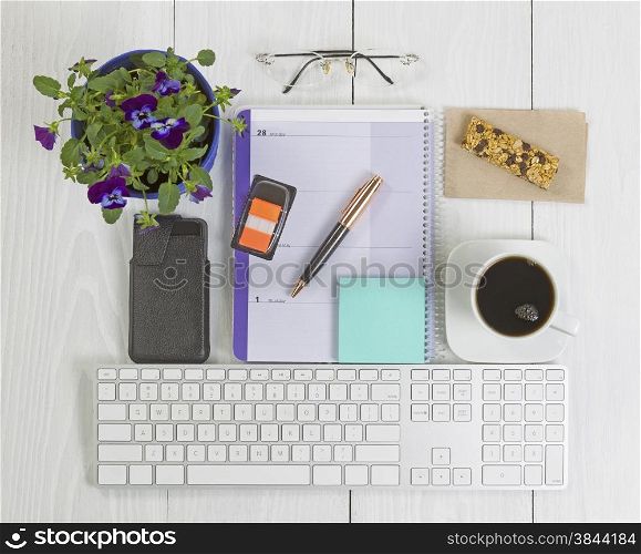 Angled top view image of a business desktop consisting of the following items: calendar, pen, reading glasses, paper stickers, house plant, keyboard and cell phone inside of case on white wood.