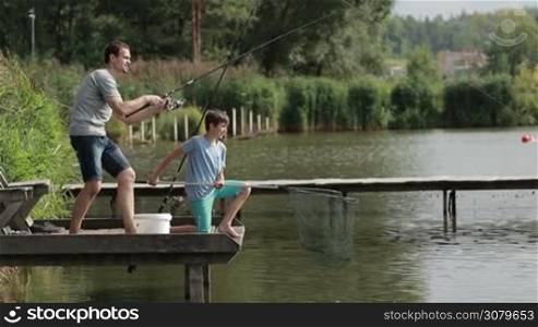 Angler with spinning rod and reel catching fish in pond while spending weekend with son on lake in countryside. Father holding rod and spining fishing reel, fighting the fish while his teenage boy with spoon fishing net helping dad to pull fish out.
