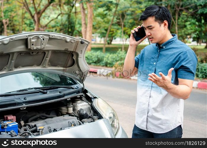 Angry Asian man and using mobile phone calling for assistance after a car breakdown on street. Concept of vehicle engine problem or accident and emergency help from Professional mechanic