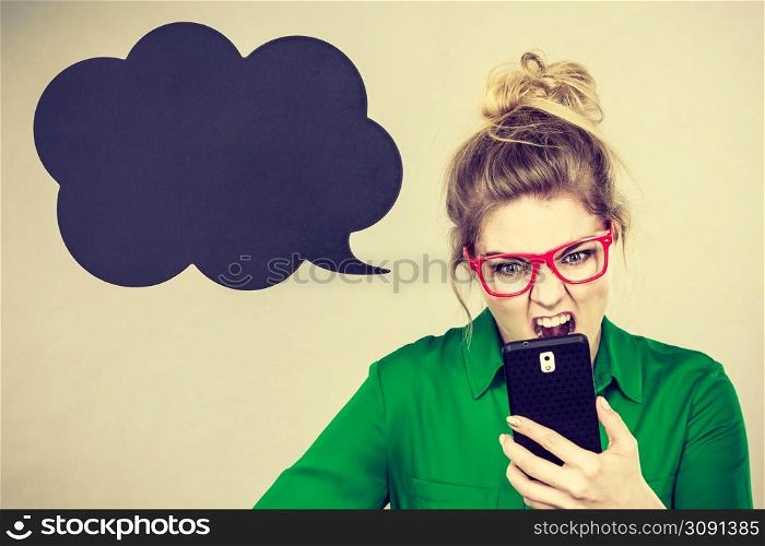 Angry business woman wearing green shirt and red eyeglasses looking at phone with black thinking or speech bubble next to her.. Angry business woman looking at phone, thinking bubble