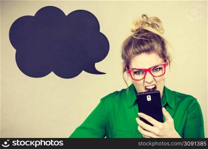 Angry business woman wearing green shirt and red eyeglasses looking at phone with black thinking or speech bubble next to her.. Angry business woman looking at phone, thinking bubble