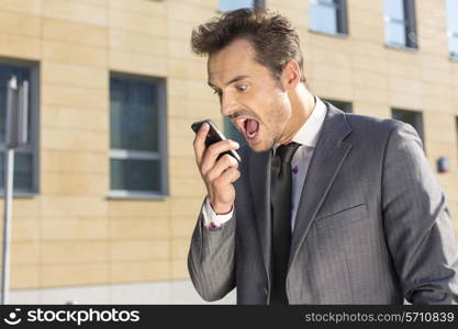 Angry businessman screaming at mobile phone against office building