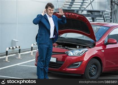 Angry businessman standing next to broken car and calling in service