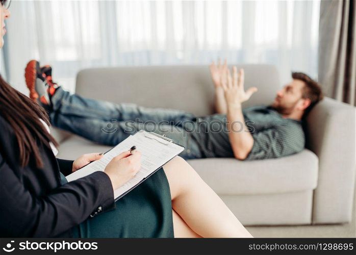 Angry male patient at psychologist reception, stress. Female doctor writes notes in notepad, professional psychology. Male patient at psychologist reception, stress