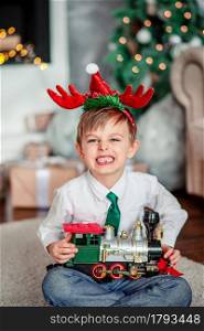 Angry upset little boy with a gift, toy train, under the Christmas tree on a New Year&rsquo;s morning. A time of miracles and fulfillment of desires. Merry Christmas.. Angry upset little boy with a gift, toy train, under the Christmas tree on New Year&rsquo;s morning. Time to fulfill wishes.