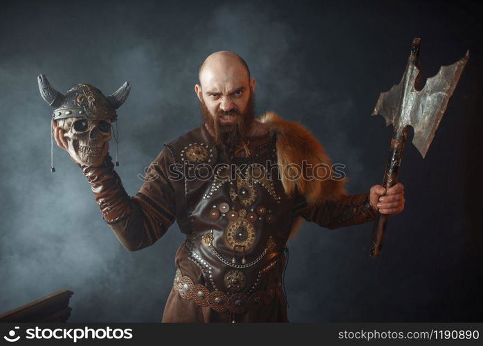 Angry viking dressed in traditional nordic clothes holds enemy&rsquo;s skull in helmet and axe, barbarian image, murderer. Ancient warrior in smoke. Angry viking holds enemy&rsquo;s skull in helmet and axe