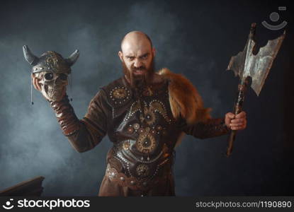 Angry viking dressed in traditional nordic clothes holds enemy&rsquo;s skull in helmet and axe, barbarian image, murderer. Ancient warrior in smoke. Angry viking holds enemy&rsquo;s skull in helmet and axe