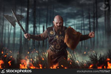 Angry viking with axe dressed in traditional nordic clothes fighting in fire, battle in forest. Scandinavian ancient warrior