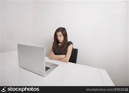 Angry young businesswoman with arms crossed looking at laptop in office