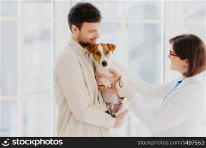 Animal care and diagnosis concept. Shot of vet, dog owner and jack russell terrier pose in veterinarian clinic, doctor examines sick animal, gives prescriptions, stand in medical office or hospital