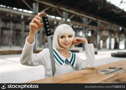 Anime style blonde lady with cold face looks at the sword. Cosplay fashion, asian culture, doll with blade, cute woman with makeup in the factory shop