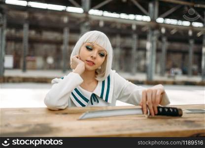 Anime style blonde lady with cold face looks at the sword. Cosplay fashion, asian culture, doll with blade, cute woman with makeup in the factory shop. Anime style lady with cold face looks at the sword