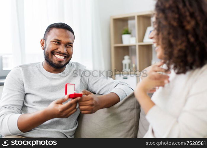 anniversary, proposal and couple concept - happy african american man giving diamond engagement ring in little red box to woman at home. african american man giving woman engagement ring