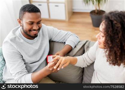 anniversary, proposal and couple concept - happy african american man giving diamond engagement ring in little red box to woman at home. african american man giving woman engagement ring
