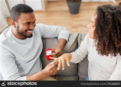 anniversary, proposal and couple concept - happy african american man giving diamond engagement ring in little red box to woman at home. african american man giving woman engagement ring