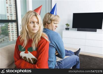 Annoyed couple in Christmas sweaters and party hats sitting back to back at home