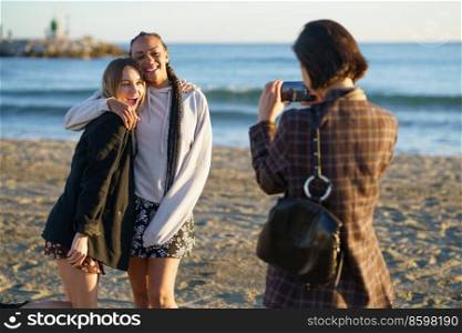 Anonymous female photographer with smartphone taking photo of cheerful multiracial female friends while standing on sandy beach near waving sea. Faceless woman photographing positive diverse girlfriends near sea