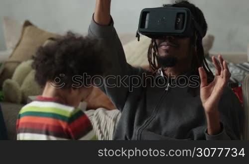 Another reality is here! Handsome young african man in VR headset sitting on the floor and gesturing in domestic interior. Hipster with dreadlocks using virtual reality goggles while mixed race son playing with toys. People using new technology.