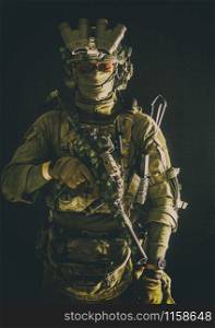 Anti-terrorist squad fighter, army elite forces soldier in combat uniform and tactical ammunition, armed mini submachine gun, wearing night-vision device, low key studio portrait on black background. Anti-terrorist squad equipped fighter soldier in darkness