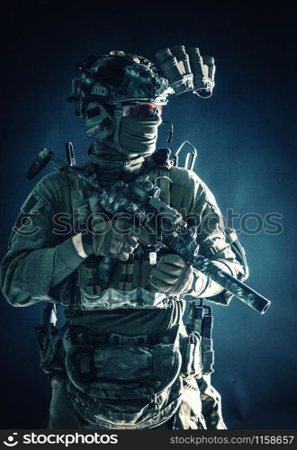 Anti-terrorist squad fighter, army elite forces soldier in combat uniform and tactical ammunition, armed mini submachine gun, wearing night-vision device, low key studio portrait on black background. Anti-terrorist squad equipped fighter soldier in darkness