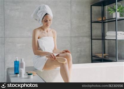 Anticellulite massage of leg with dry brush. Beautiful happy lady is making body massage with brush in bathroom. Attractive caucasian woman wrapped in towel after bathing. Girl takes shower at home.. Anticellulite massage with dry brush. Attractive caucasian woman wrapped in towel after bathing.