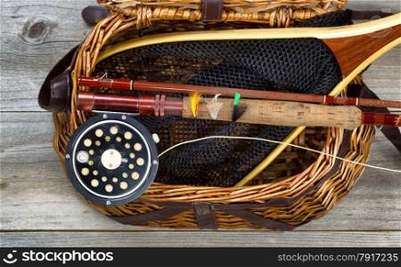 Antique fly fishing reel, rod, flies, and net on top of open creel with rustic wood underneath. Layout in horizontal format.